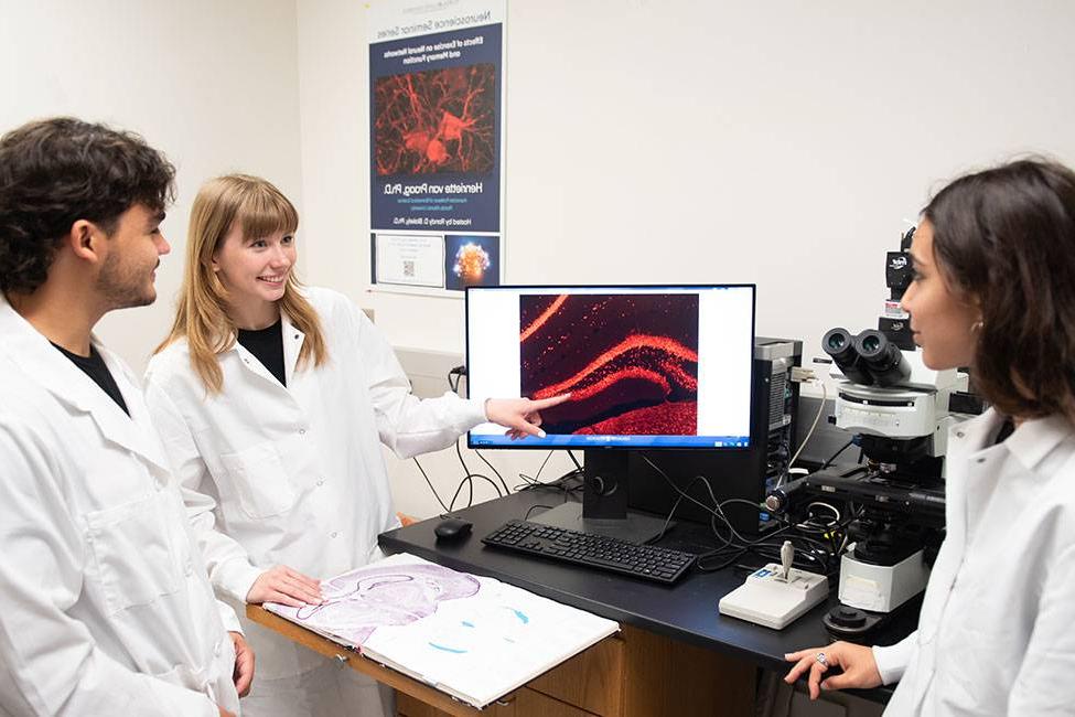 Three scientists wearing white lab coats and standing around a computer screen. The screen shows a microscope image. One female is pointing to the image while the others look on.